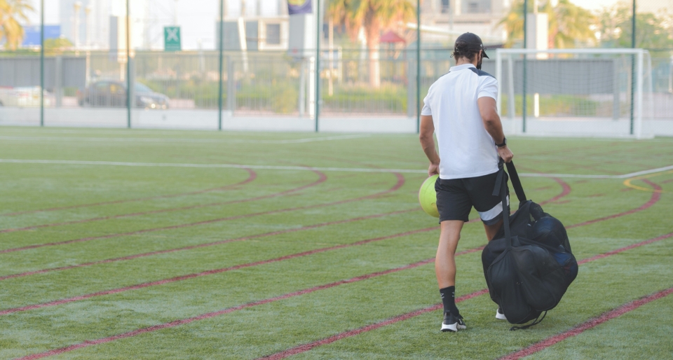 A football coach carrying a bag of footballs across a pitch